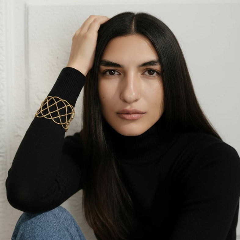 Woman wearing a black turtleneck and blue jeans with a gold arm band, sitting against a white wall.
