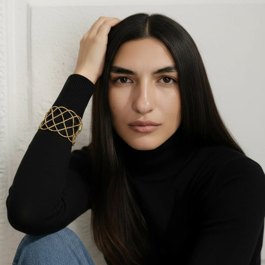 Woman wearing a black turtleneck and blue jeans with a gold arm band, sitting against a white wall.