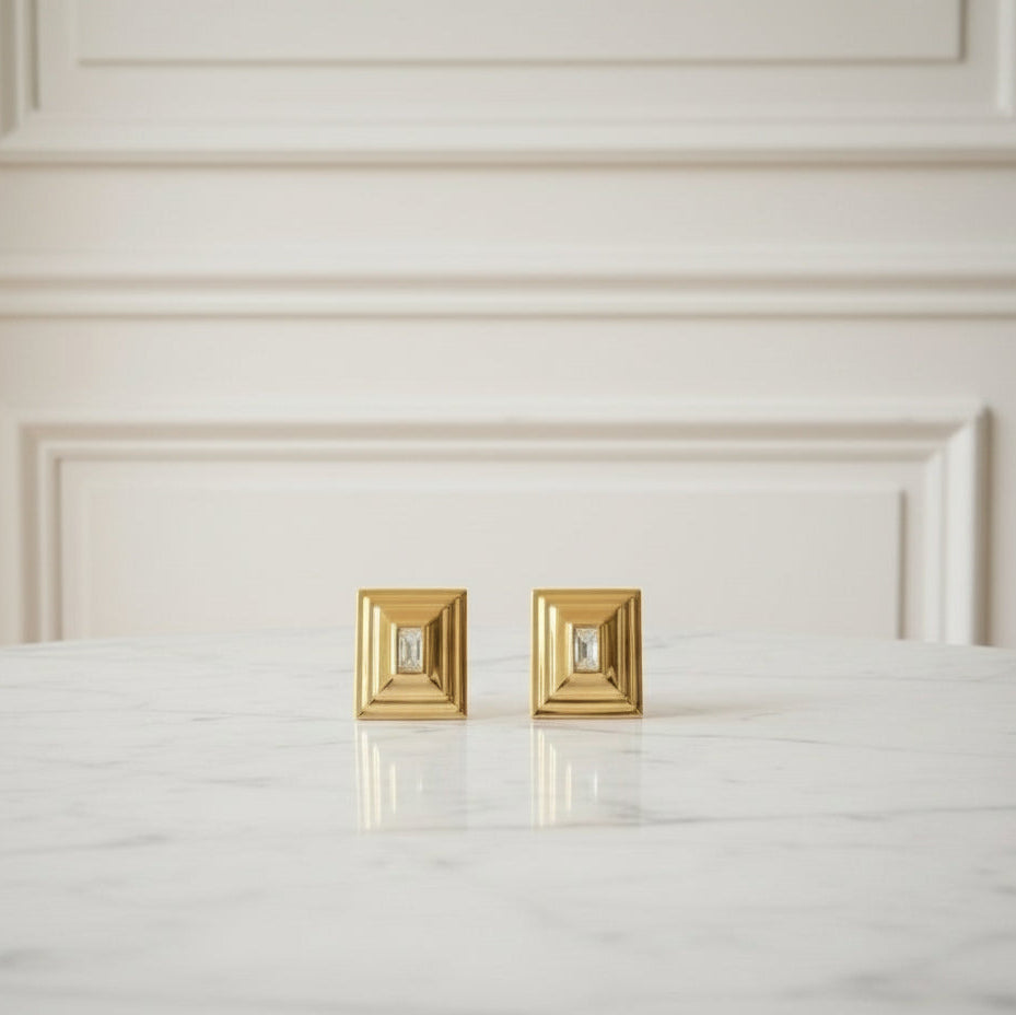 Gold square earrings on a white surface with a neutral background